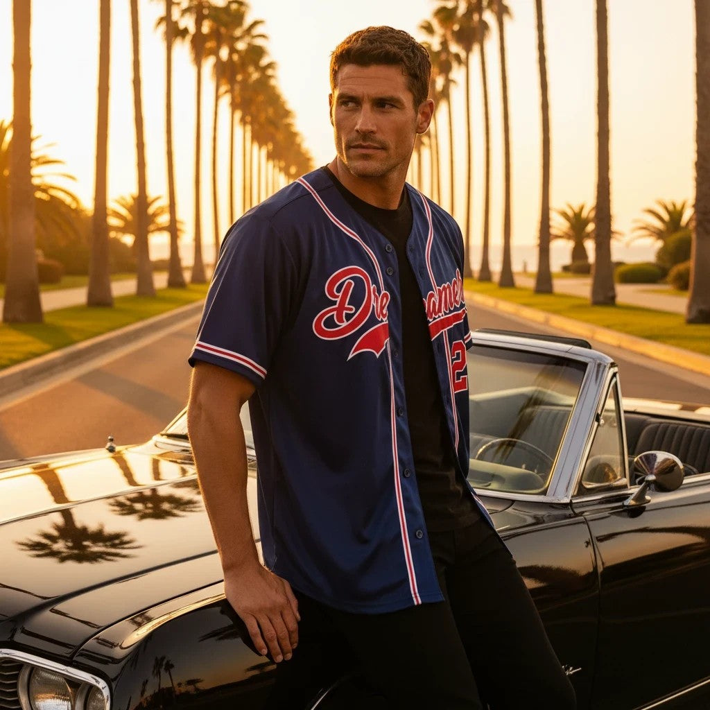Man wearing the royal-blue Dreamer Baseball Jersey beside a black classic car on a warm palm-lined coastal road.